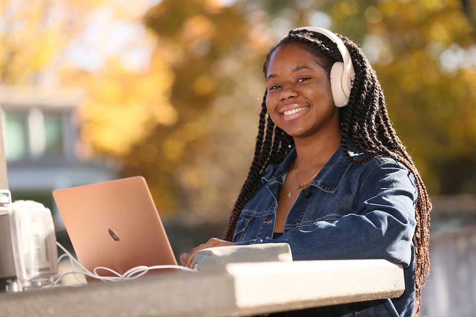Female student wearing headphones in front of laptop smiles at camera.