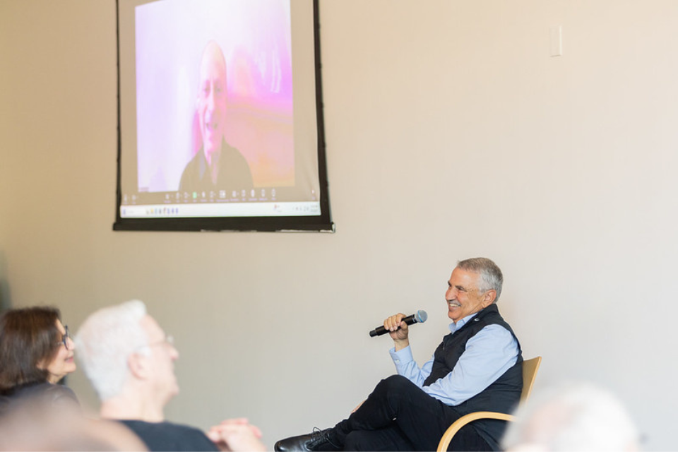 Man seated with microphone speaking to a crowd with another man on screen to the left of him