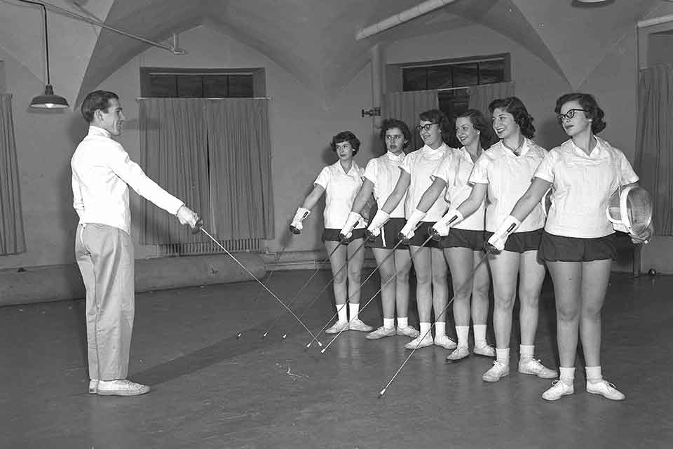 Women standing in a line with fencing foils being taught by a male instructor.