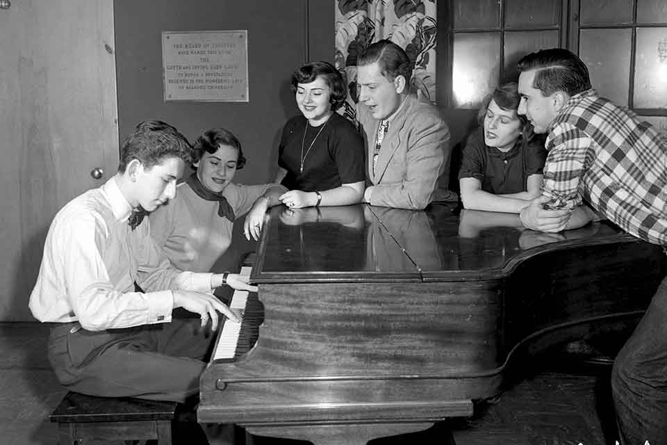 Group of students standing around a piano as a male student plays
