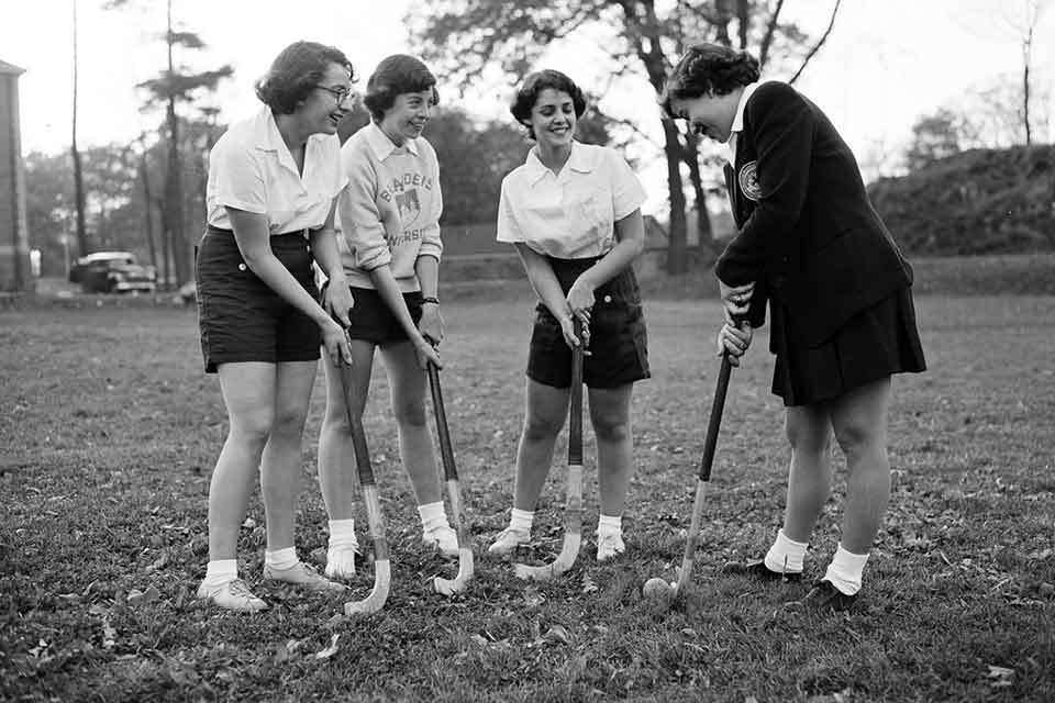 Group of Brandeis women in uniform standing in a circle with field hockey sticks