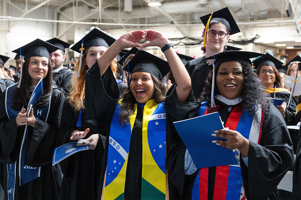 Group of female students wearing caps and gowns at commencement. One is making a heart with her hands.