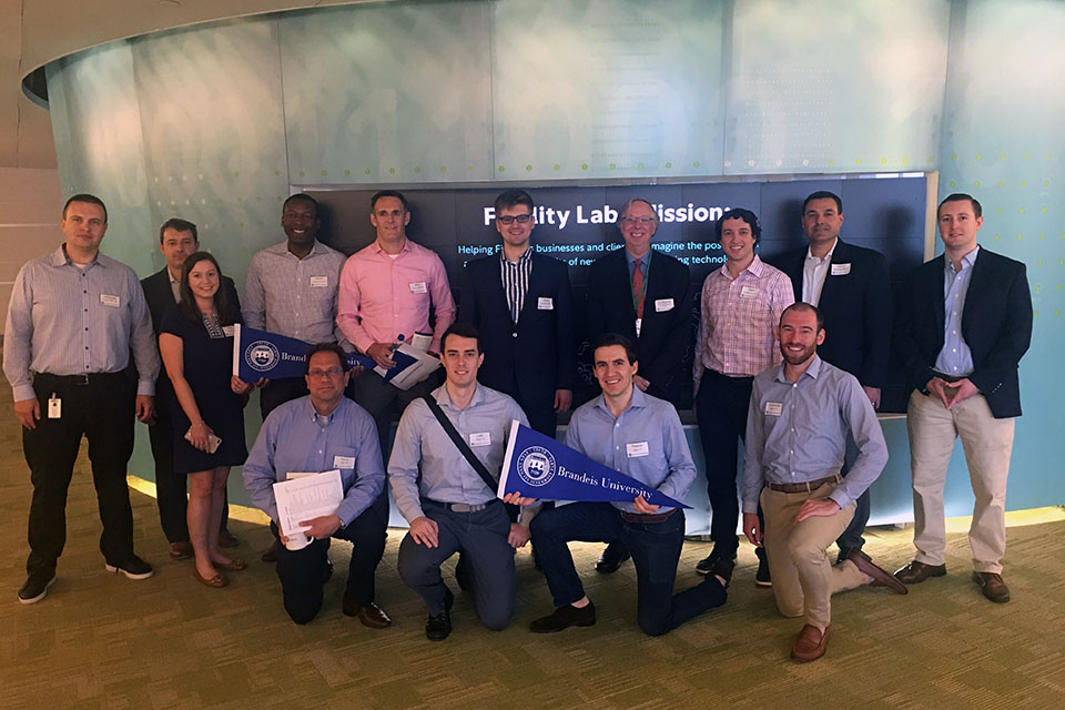 A group of alumni holding Brandeis pennants stand in front of wall with Fidelity logo