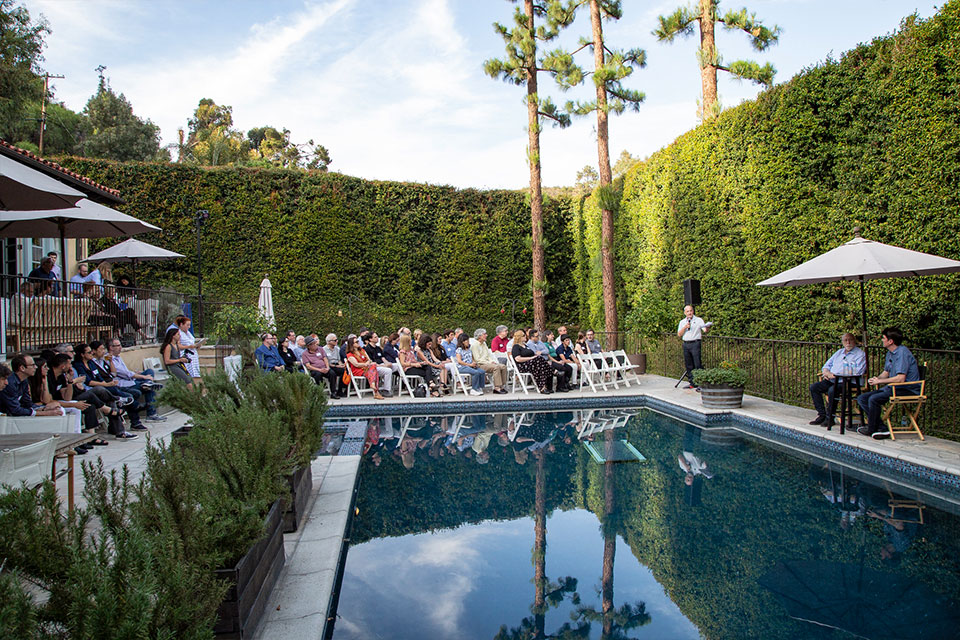 A seated crowd surrounds a swimming pool and looks at speakers with microphones