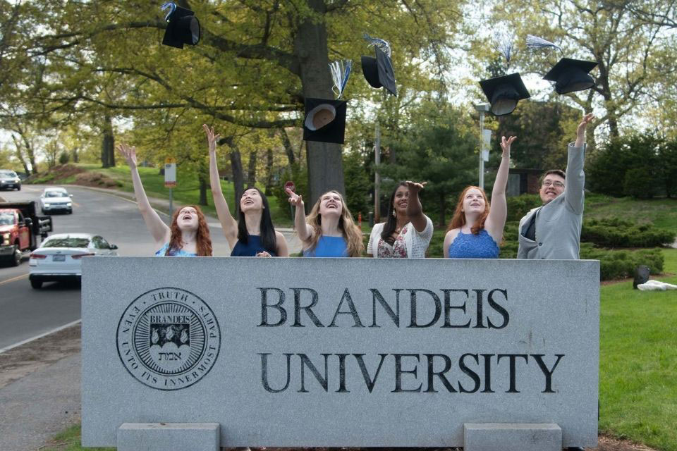students tossing graduation caps behind Brandeis sign 