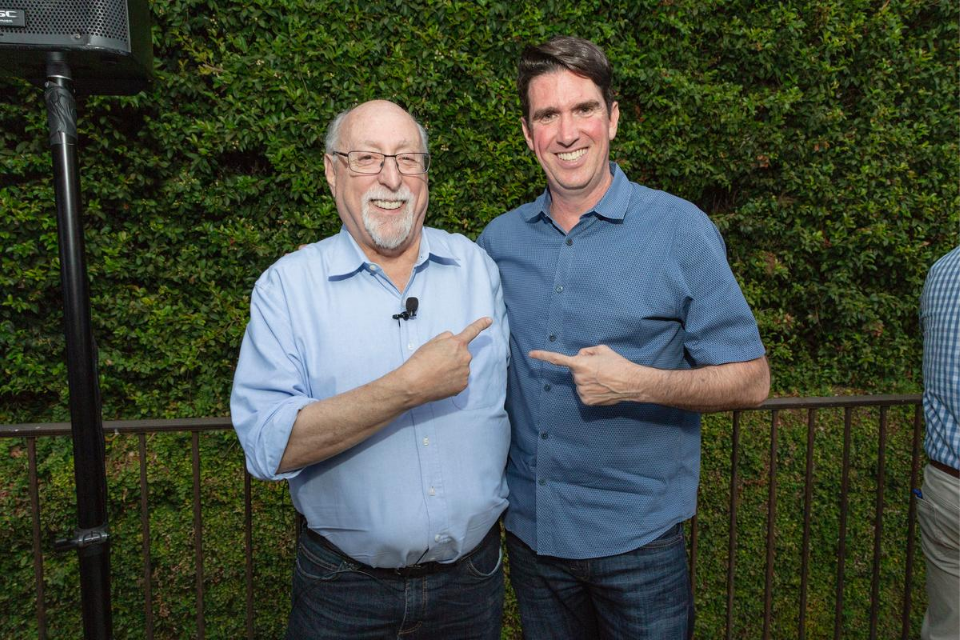 Walt Mossberg and Adam Cheyer smile widely at the camera and point fingers at one another in front of greenery