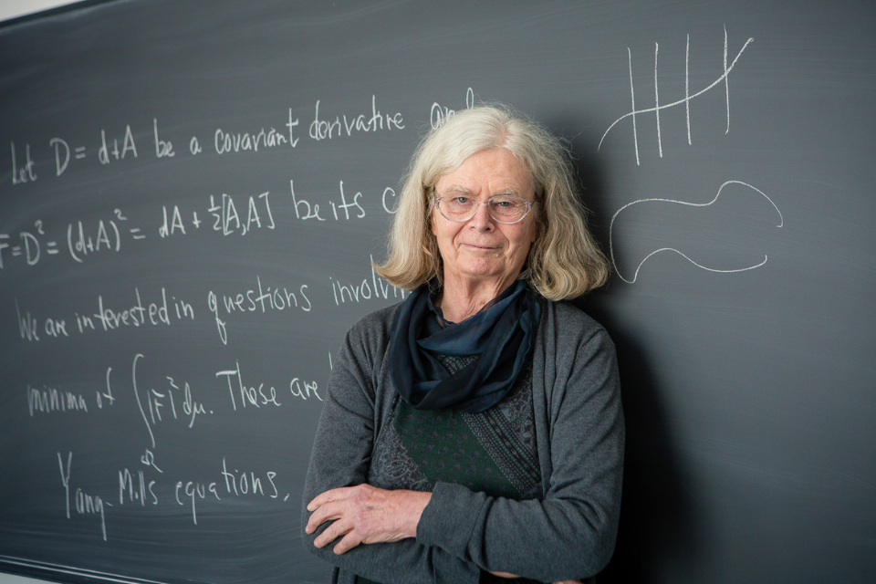 Karen Uhlenbeck stands with her arms crossed in front of a chalkboard with mathematical equations on it