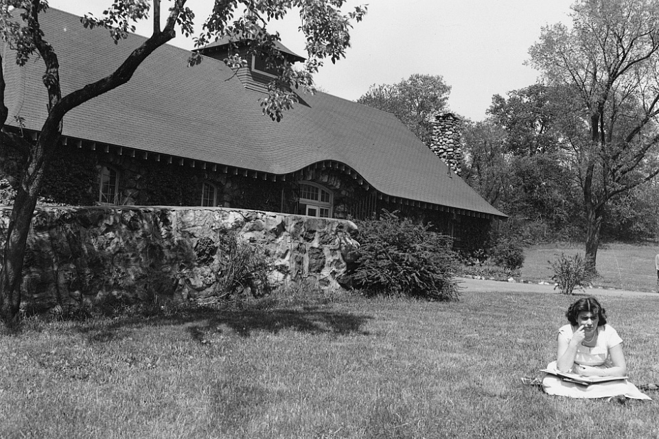 A woman sits with a book in her lap in a field in front of a stone building