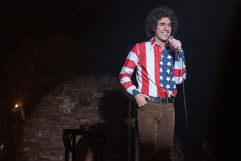 Sasha Baron Cohen, as Abbie Hoffman, standing with a microphone and wearing an American flag shirt