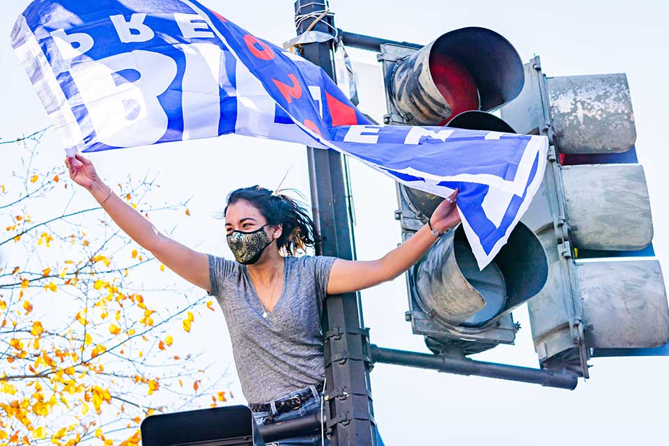 Woman with mask on sitting on top of a traffic light waving a Biden for president flag