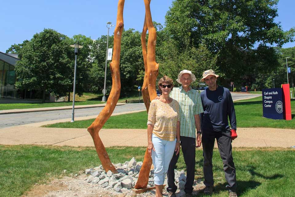 Madeline Calabrese, Harold Grinspoon and Adam Suska stand by a wooden sculpture on the Brandeis campus. 