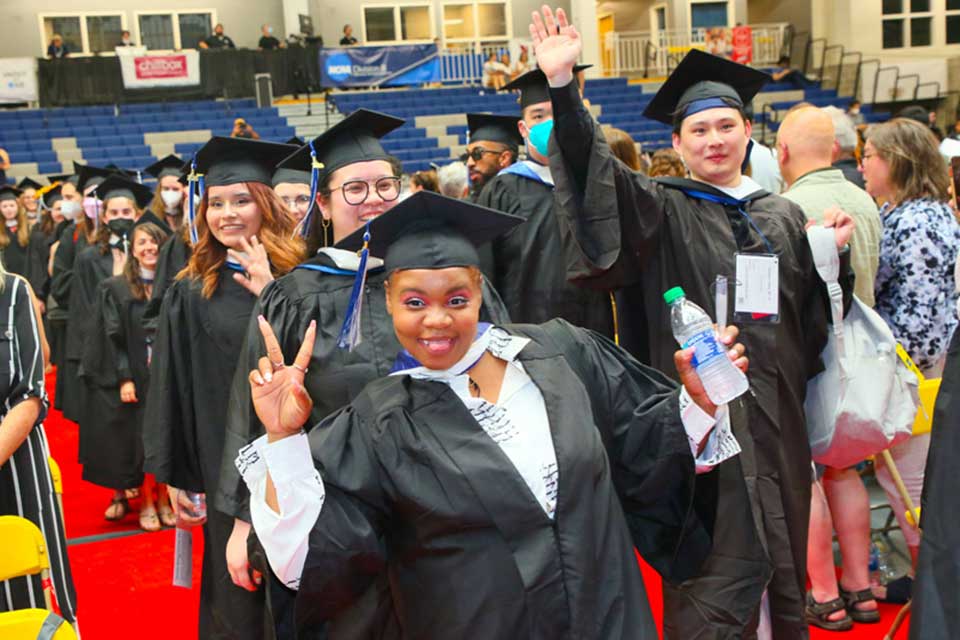 Female graduate gives peace sign and smiles in front of a line of graduates 