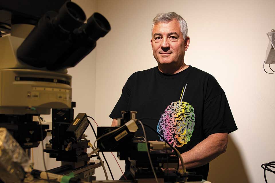 Man with gray hair and black t-shirt with a brain on it stands next to some lab equipment.