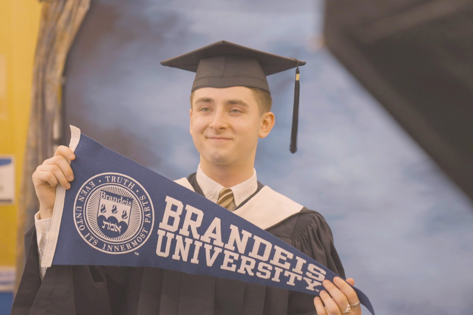 Brandeis graduate in cap and gown holds up a pennant