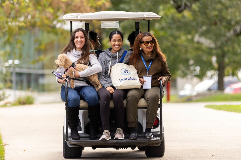 Three women and a small dog smiling and riding on the back of a  golf cart.