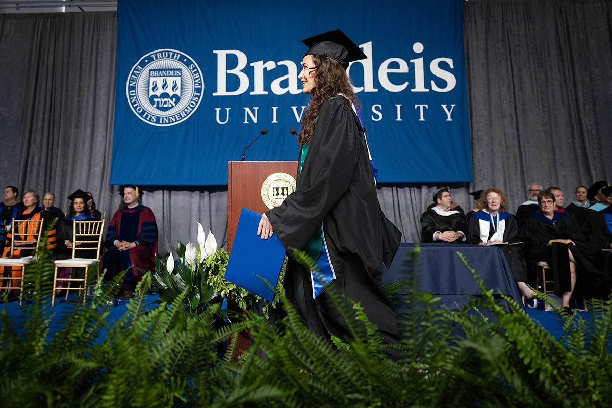 A graduate walks across the stage during Commencement. A Brandeis banner is seen in the background.
