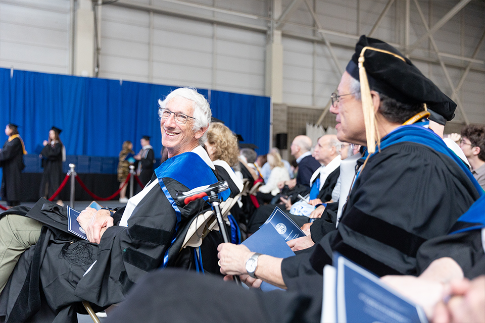 A man turned around seated at a commencement proceedings, smiling in academic regalia.