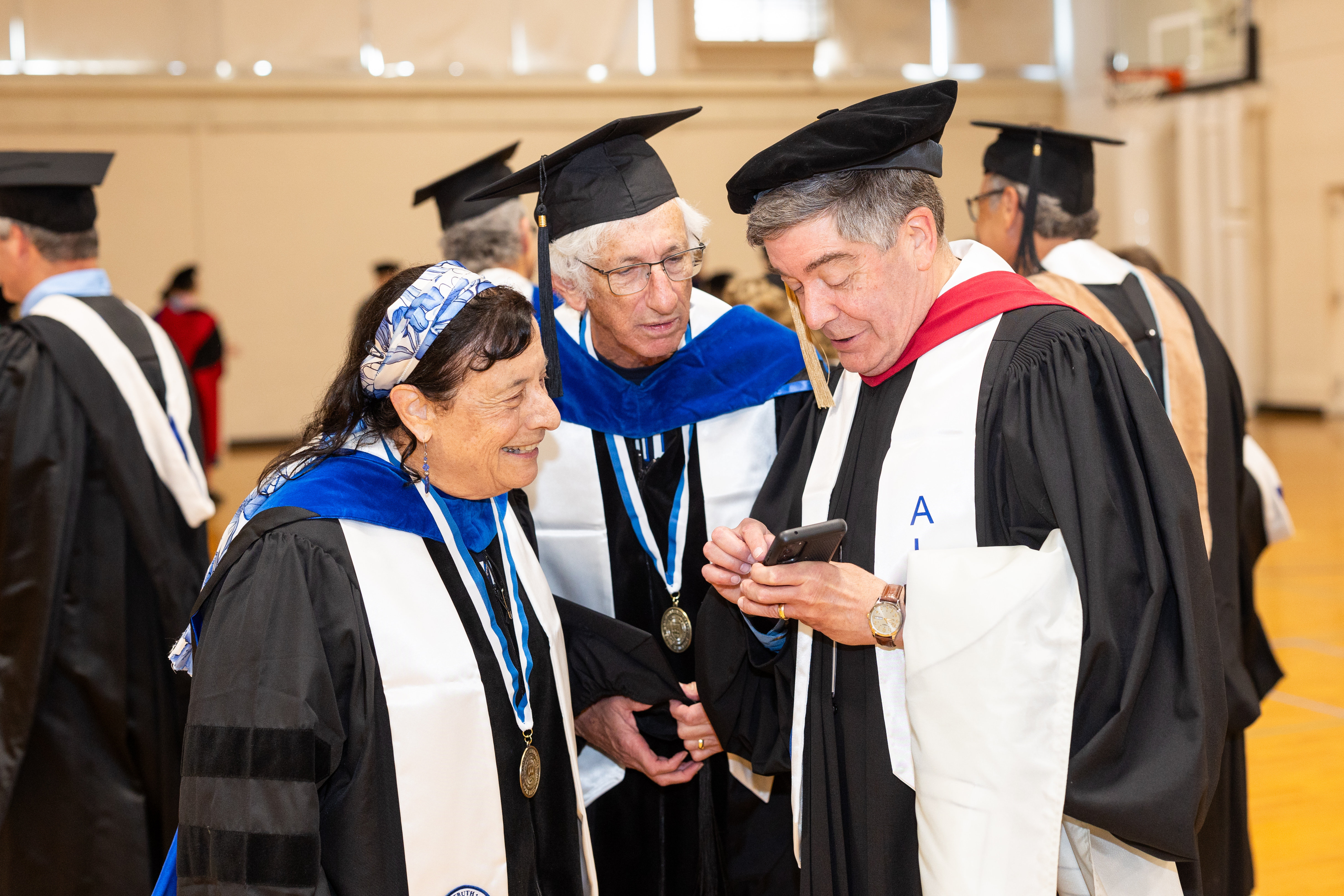 A group of alumni from the class of 1975 stand robed and smiling at each other looking at a phone sharing a memory together