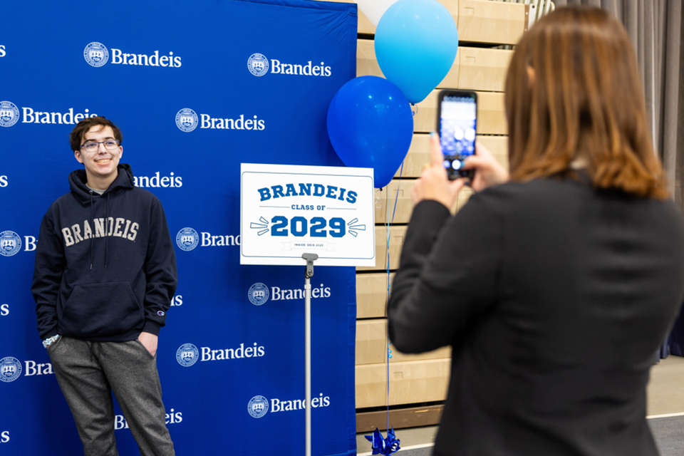 Student smiling in front of Brandeis step and repeat backdrop having his photo taken by balloons and "class of 2029" sign