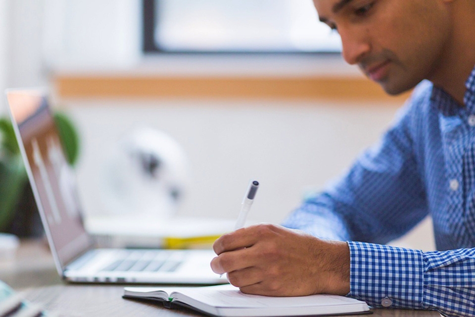 Person sits at a computer while writing something in a notebook