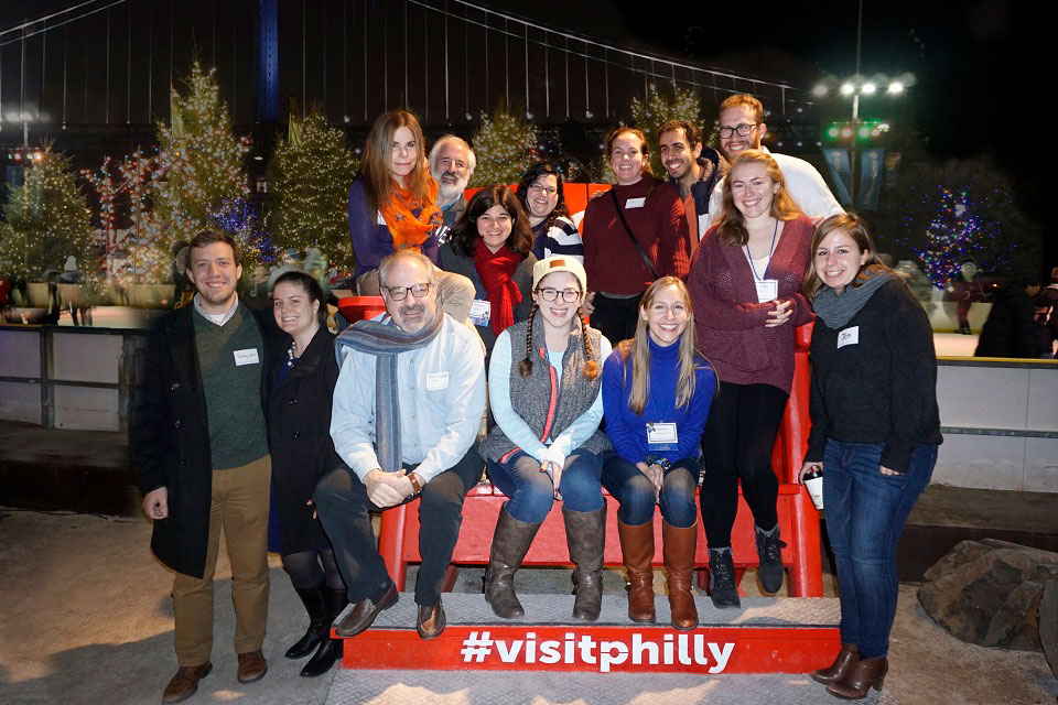 Attendees at the holiday gathering pose for a photo in a large chair with the Delaware River and Ben Franklin Bridge in the background in December 2018.