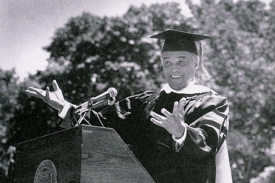 Harry Belafonte with cap and gown smiling at lectern with hands up