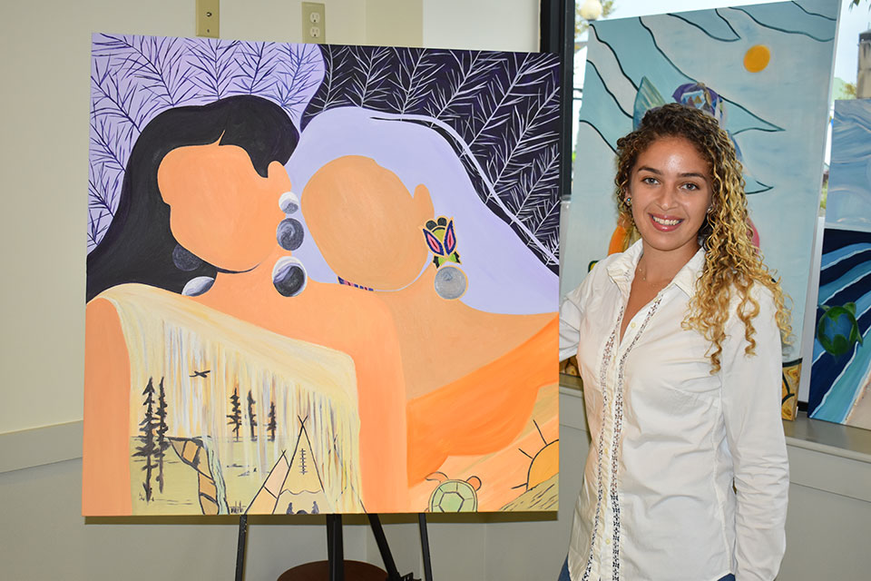 Natasha Frye standing next to one of her paintings of two Indigenous women.