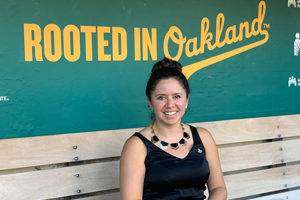 Amelia Schimmel smiling and seated on a bench in front of a slogan that reads, "Rooted in Oakland". 