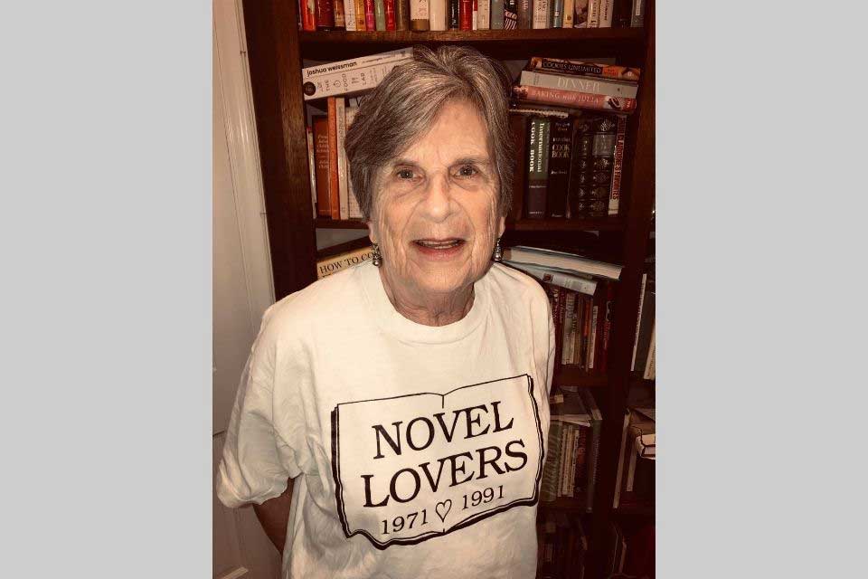 Elinor Berlin standing in front of a book case wearing a t-shirt that says: Novel Lovers 1971 1981