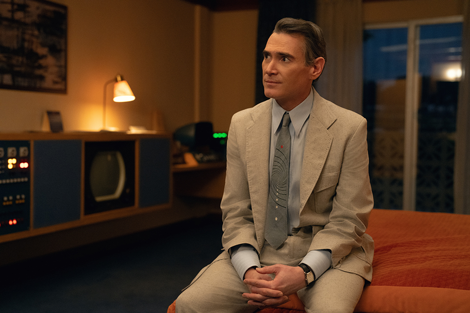 Hello Tomorrow! star Billy Crudup sits on a bed at the Vista Motor Lodge hotel room with a mix of retro and futuristic technology behind him, including a 1950s-style television.