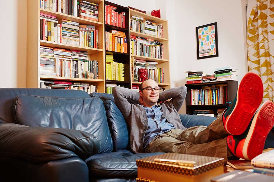 Josh Gondelman sitting on a couch with books on bookshelves in the background. 